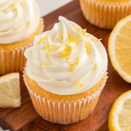 Recipe card image showing Lemon Cream Cheese Frosting piped high on a lemon cupcake with fresh lemon zest and sliced lemons on a wooden board.