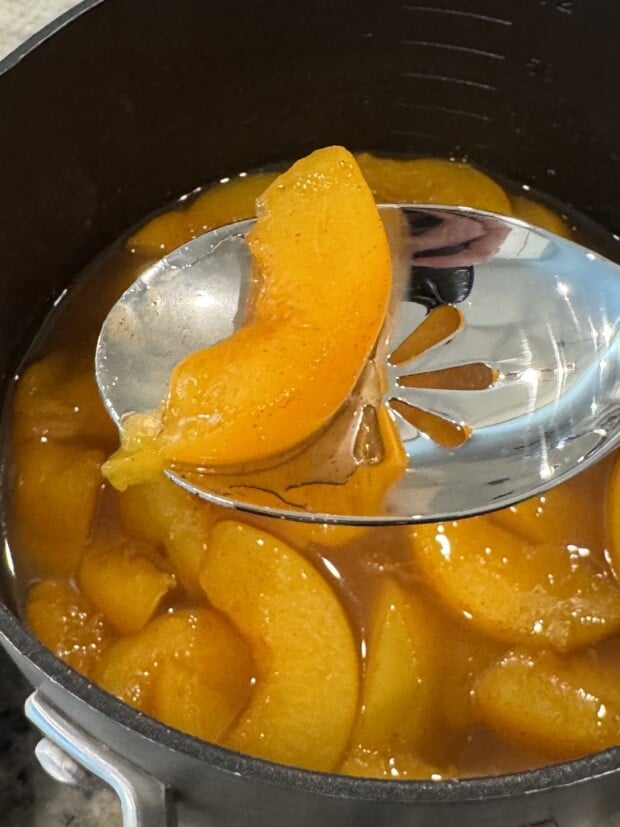 Slotted spoon lifting a cooked peach slice from spiced syrup to drain excess liquid