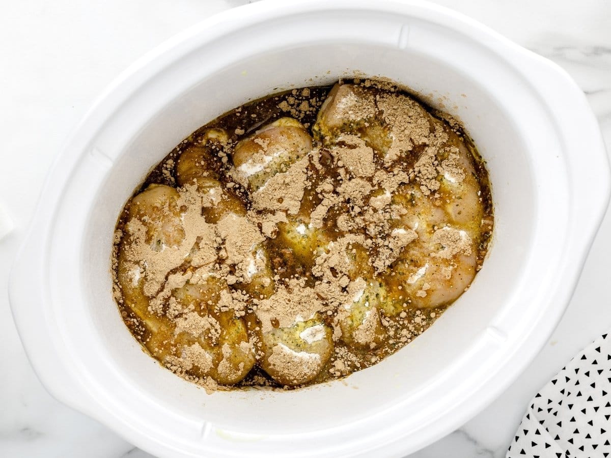 Chicken breasts in a slow cooker topped with ranch seasoning, gravy mix, butter, and pepperoncini juice before cooking, showing the layered ingredients ready to cook.
