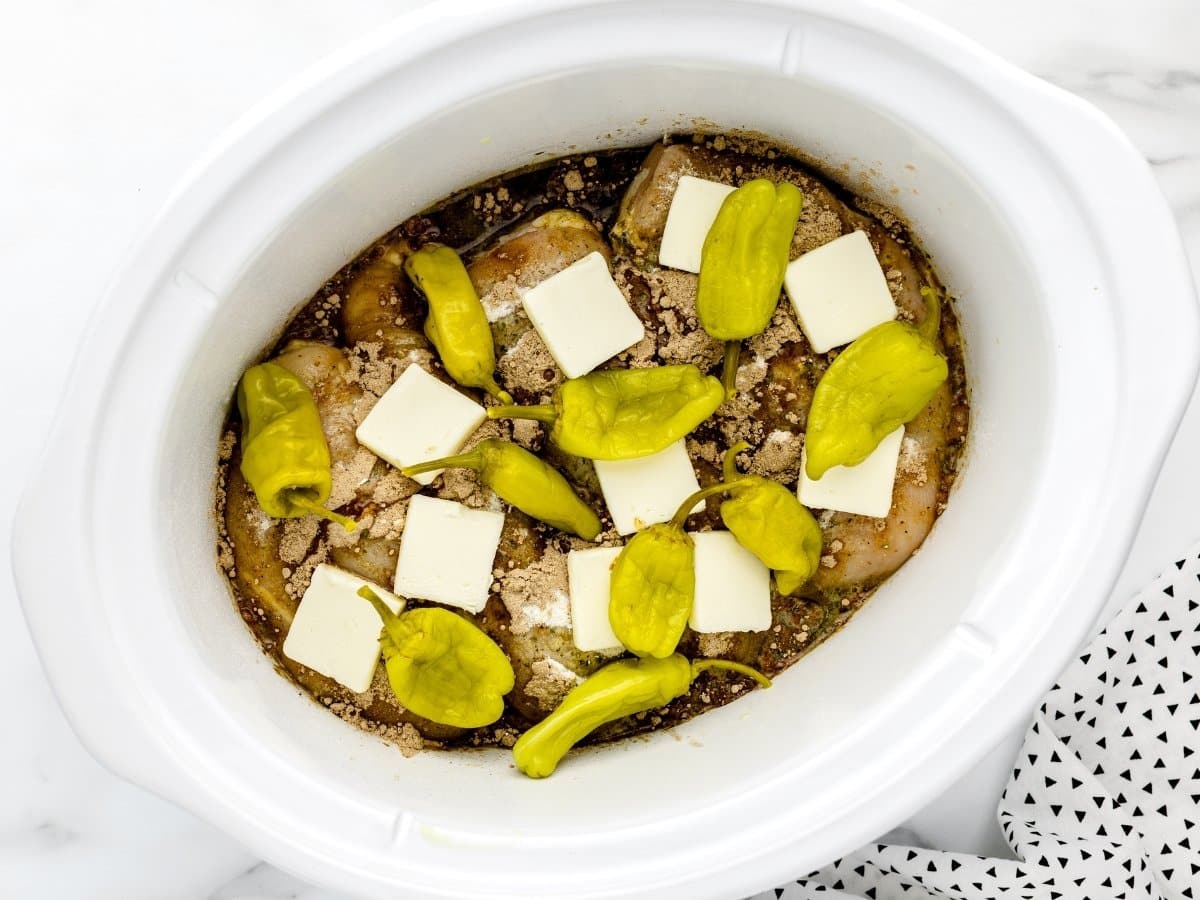 Chicken breasts in a slow cooker topped with ranch seasoning, gravy mix, sliced butter, and whole pepperoncini peppers before cooking, showing the ingredients layered and ready to cook.