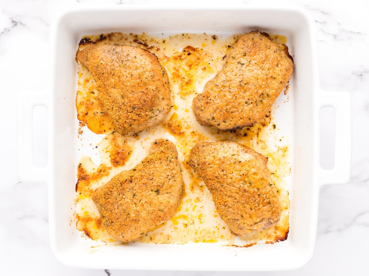 Overhead view of four baked Parmesan crusted pork chops in a white casserole dish, with a golden breadcrumb coating and browned edges, surrounded by melted cheese and juices.