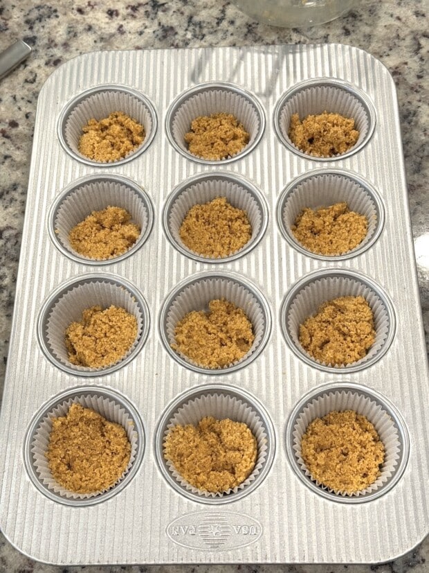 Graham cracker crust mixture portioned into lined muffin pan cups before pressing