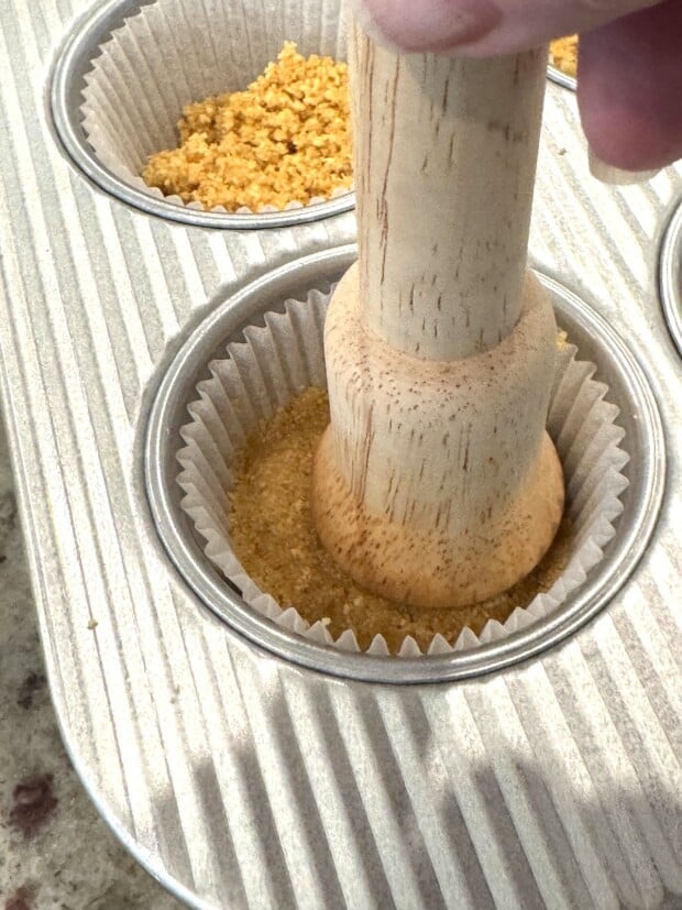Pressing graham cracker crumbs firmly into muffin liner using a wooden tart press