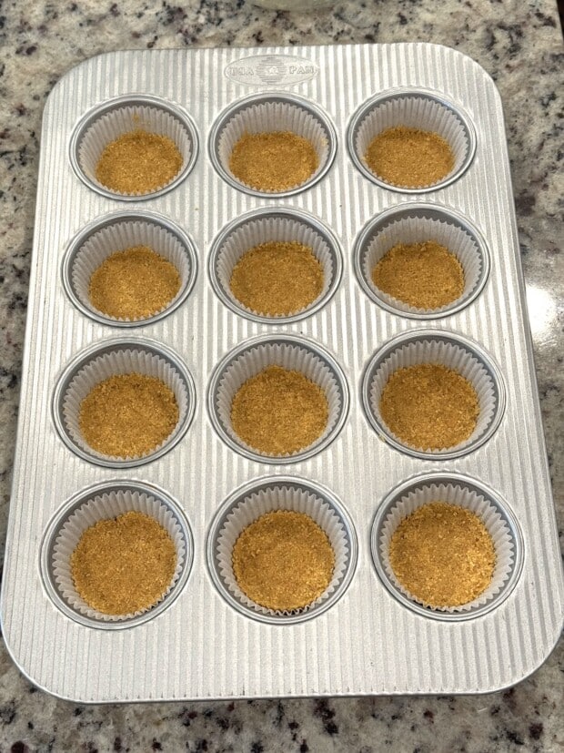 Evenly pressed graham cracker crusts in a lined muffin pan ready for baking