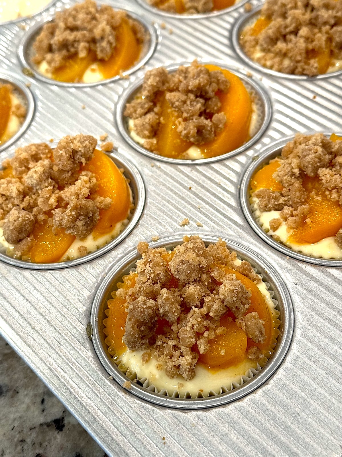 Mini peach cobbler cheesecakes topped with crumble before baking in muffin pan