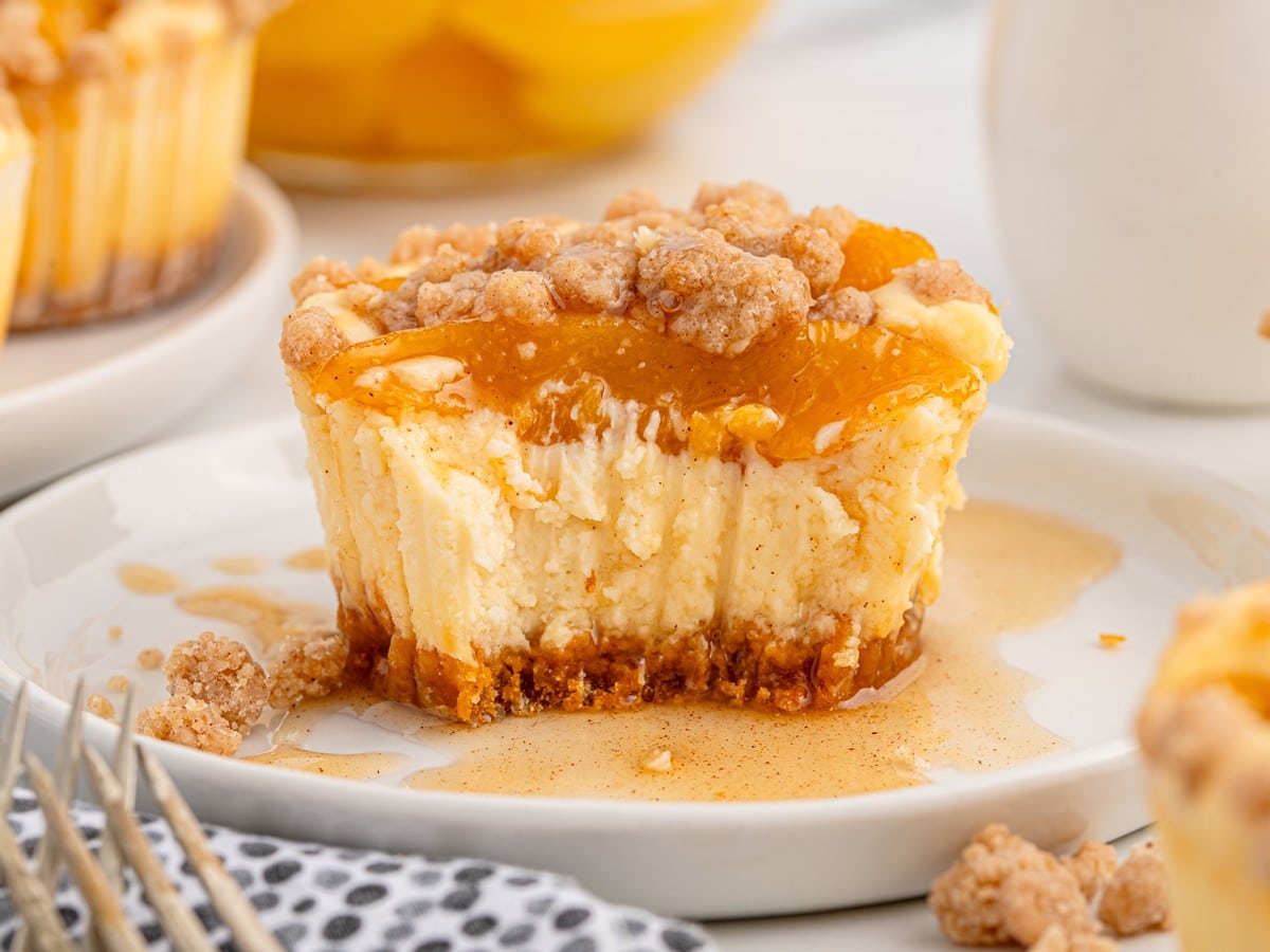 Single peach cobbler cheesecake bite with a bite taken out, showing creamy cheesecake texture, peach topping, crumbly streusel, and graham crust on a white plate with glaze drizzle