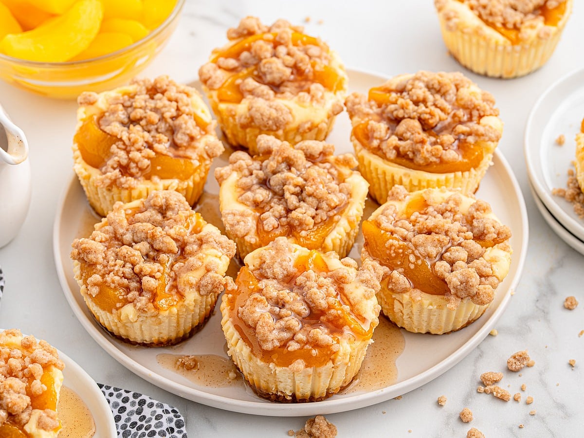 Overhead view of multiple peach cobbler cheesecake bites on a white plate, topped with peach filling and cinnamon crumble, surrounded by crumbs and glaze on a light surface