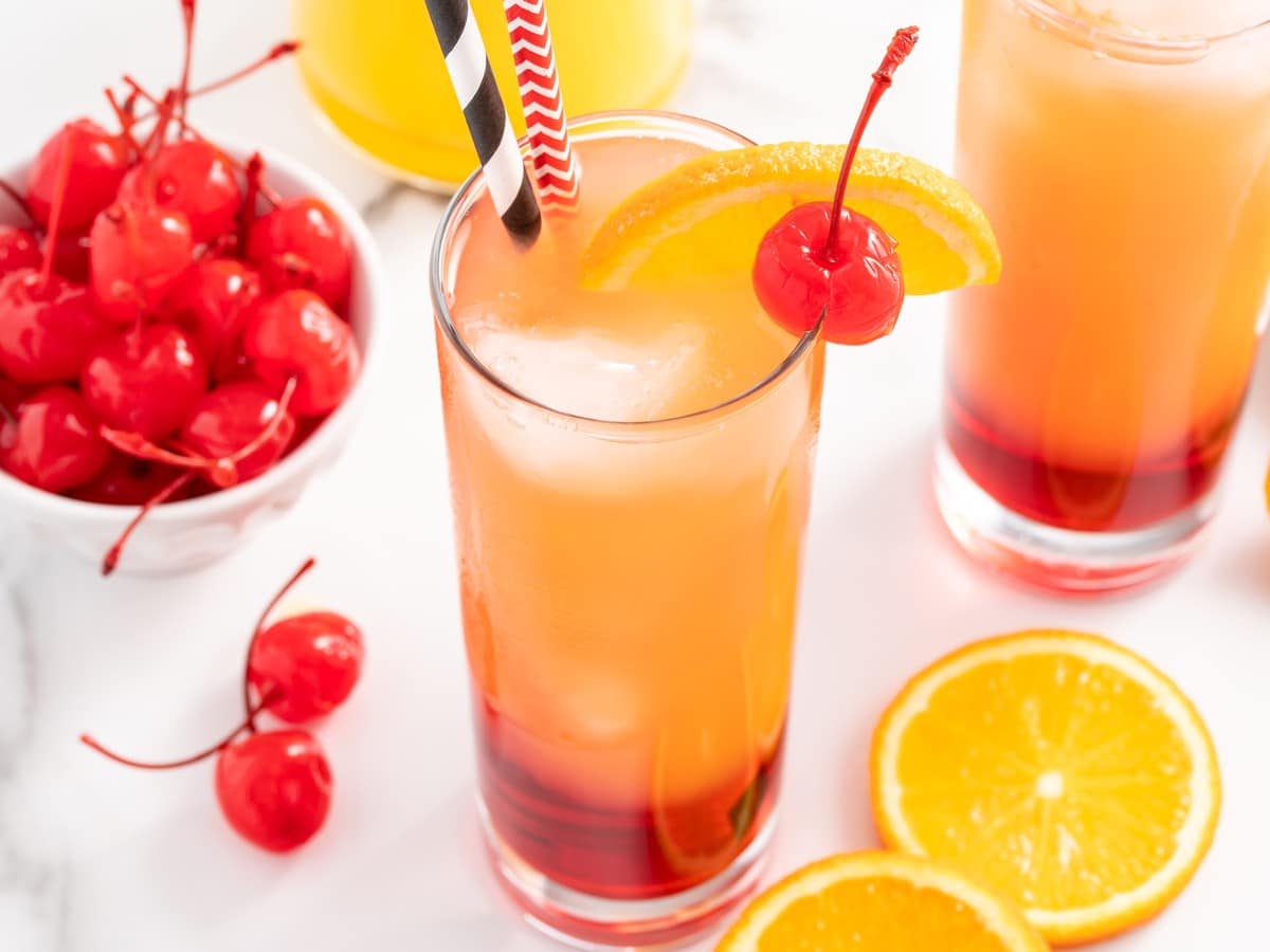 Close-up of a Sex on the Beach cocktail with ice, orange slice, maraschino cherry, striped straws, and red grenadine layer at the bottom.