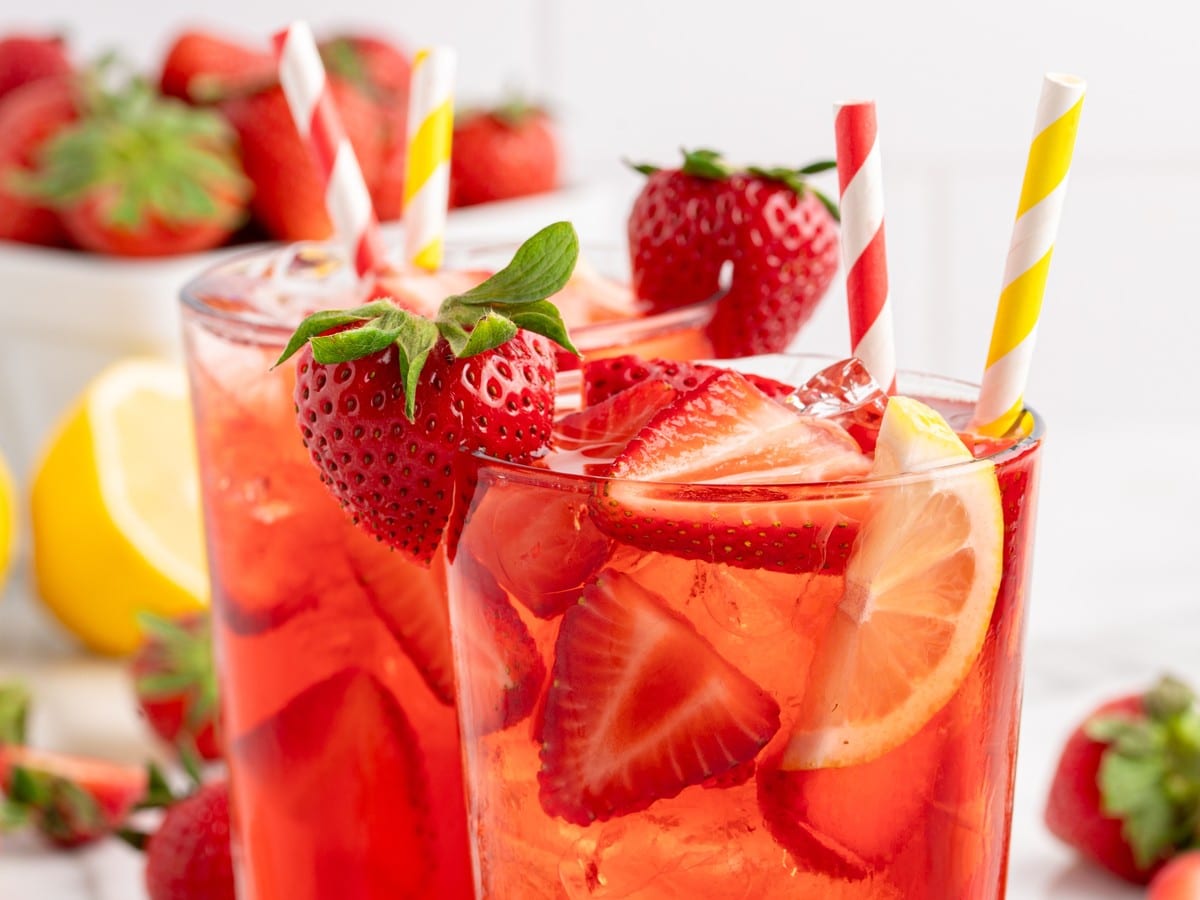Close-up of Southern Strawberry Sweet Tea in tall glasses with ice, fresh strawberry garnish, lemon slices, and striped paper straws.
