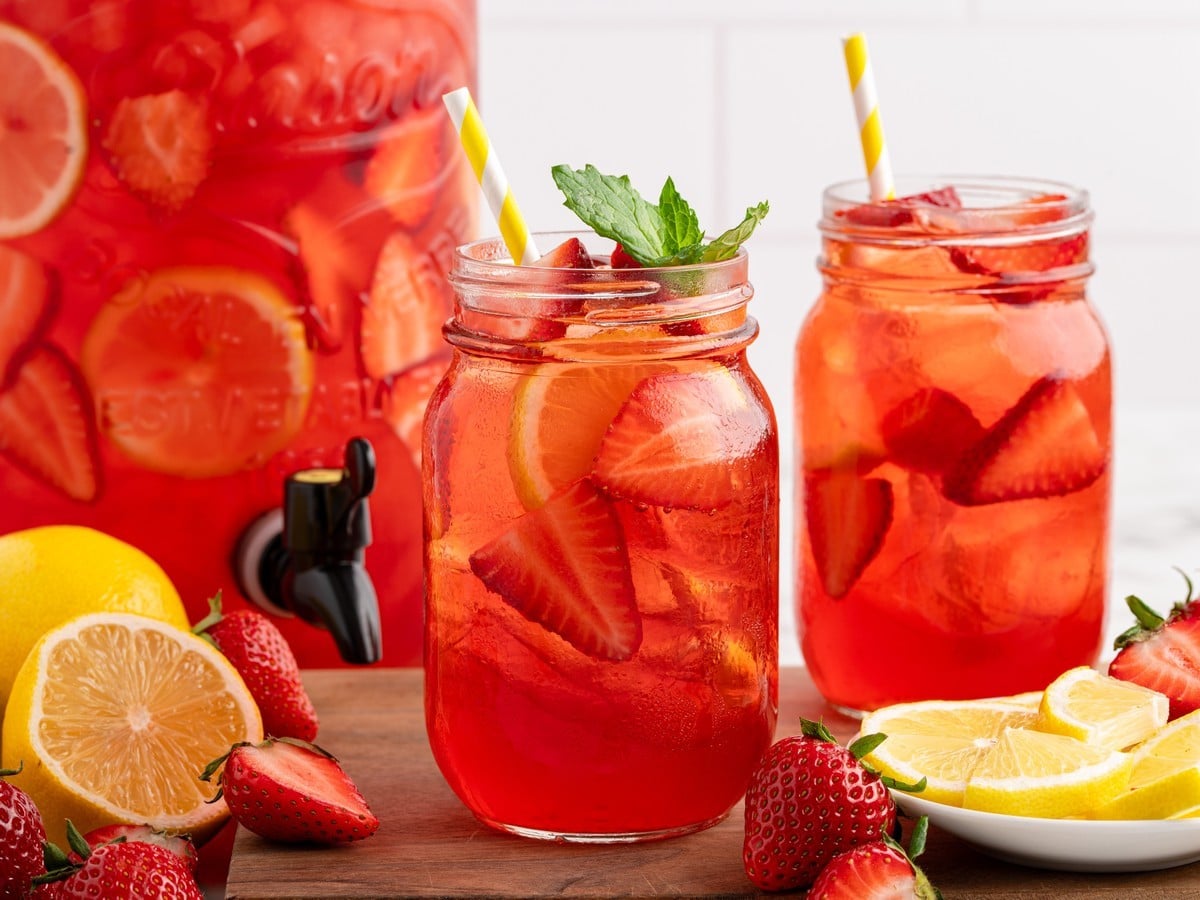 Mason jar glasses of Southern Strawberry Sweet Tea filled with ice, sliced strawberries, lemon slices, and mint, served beside a drink dispenser with fresh strawberries and lemons.