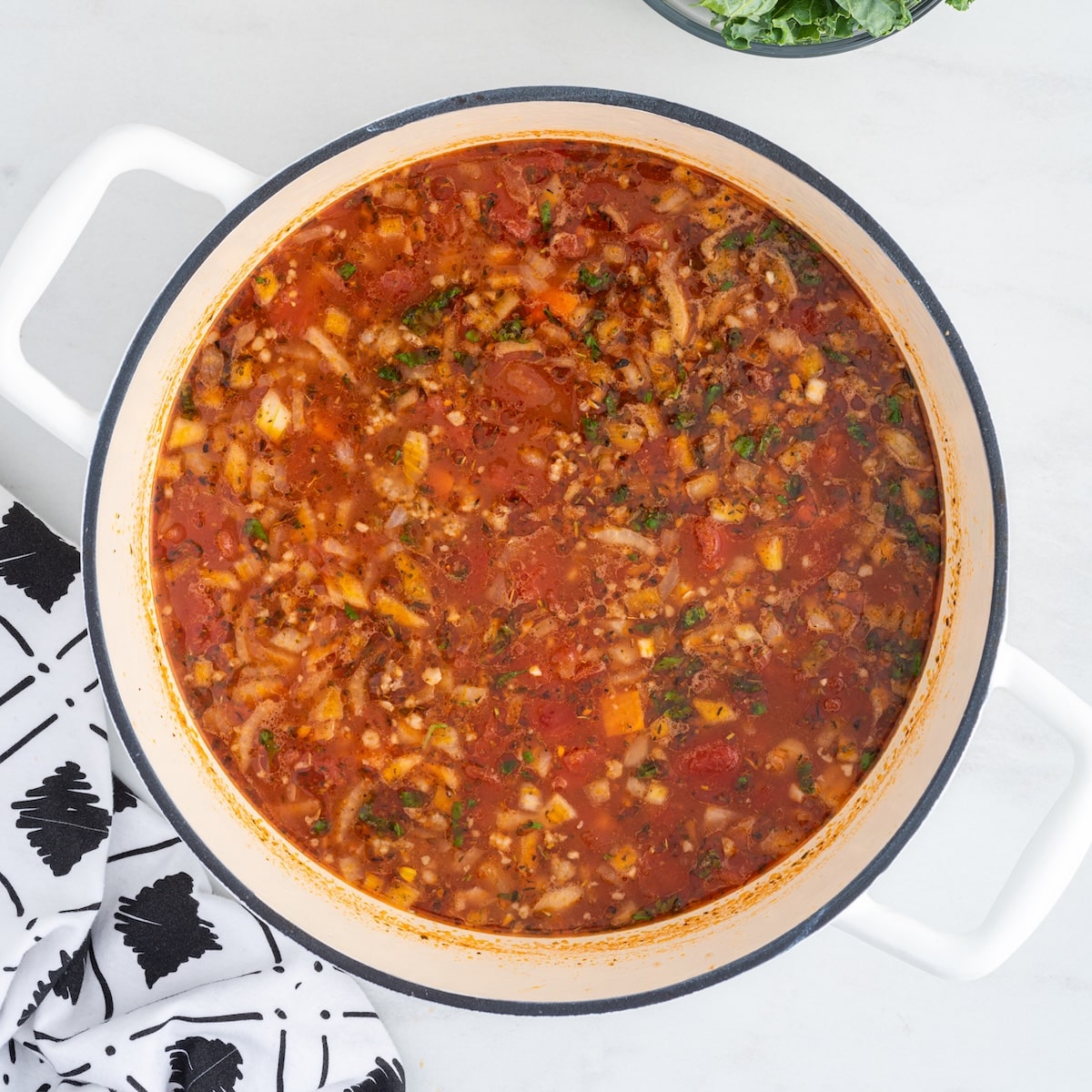 A white Dutch oven filled with simmered sausage tortellini soup broth, with bowls of fresh cheese tortellini and chopped kale ready to be added.