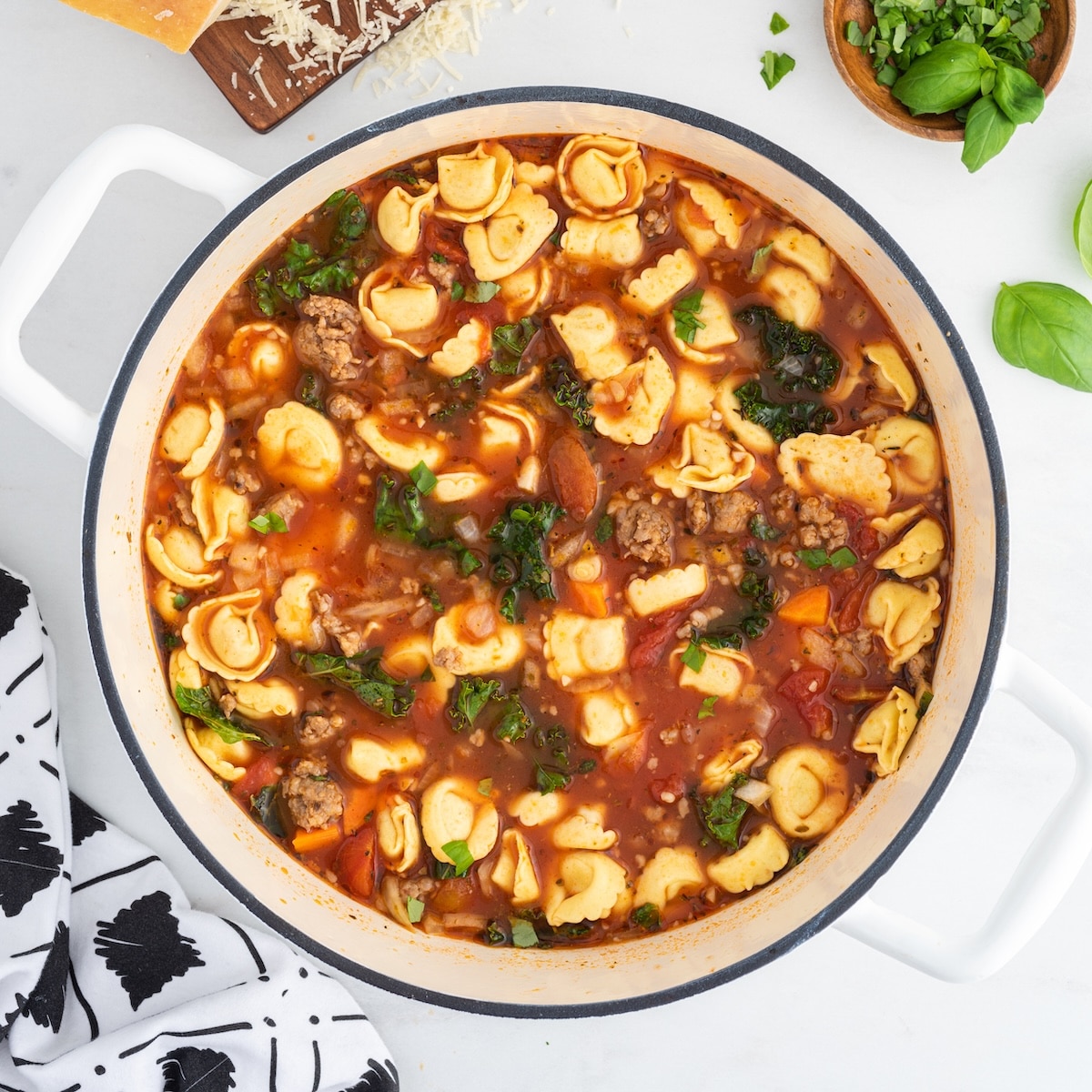 A white Dutch oven filled with finished sausage tortellini soup with kale, surrounded by a block of parmesan on a grater, fresh chopped basil, and soup ladles.