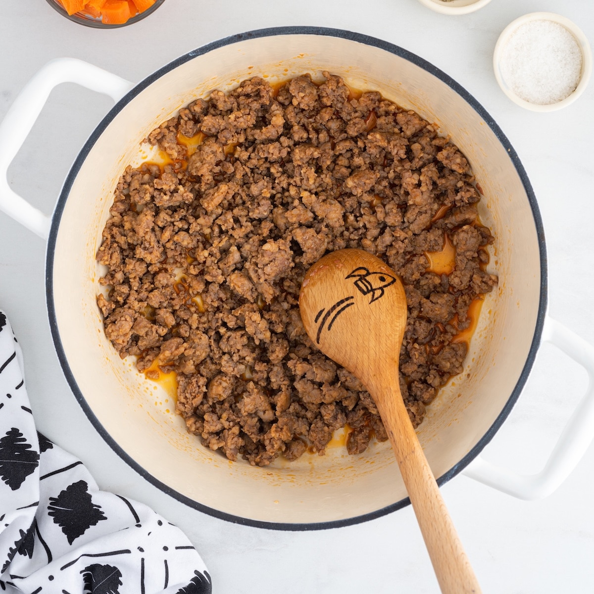 Ground Italian sausage browned in a white Dutch oven with a wooden spoon, with diced carrots and a bowl of salt visible in the background.