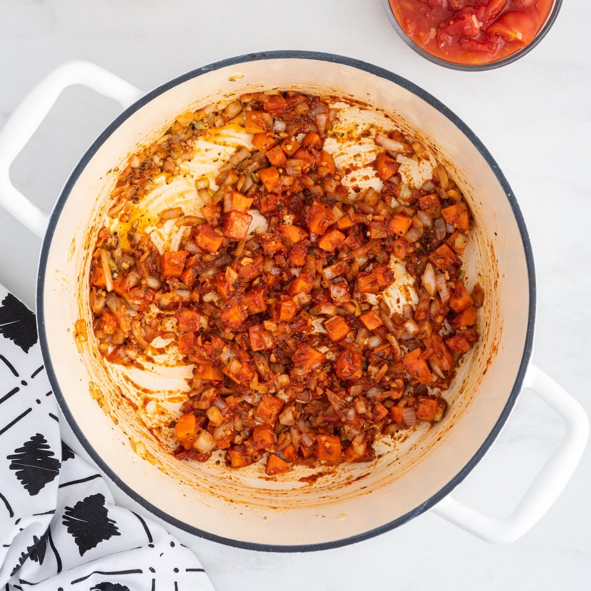 Diced carrots, onion, and tomato paste cooked together in a white Dutch oven, with a bowl of stewed tomatoes visible in the background.