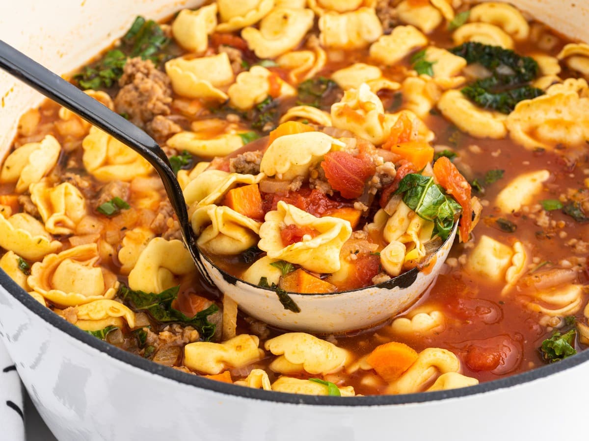 Close-up of tortellini soup in a white Dutch oven with a black ladle scooping tortellini, sausage, tomatoes, carrots, and kale in rich tomato broth.