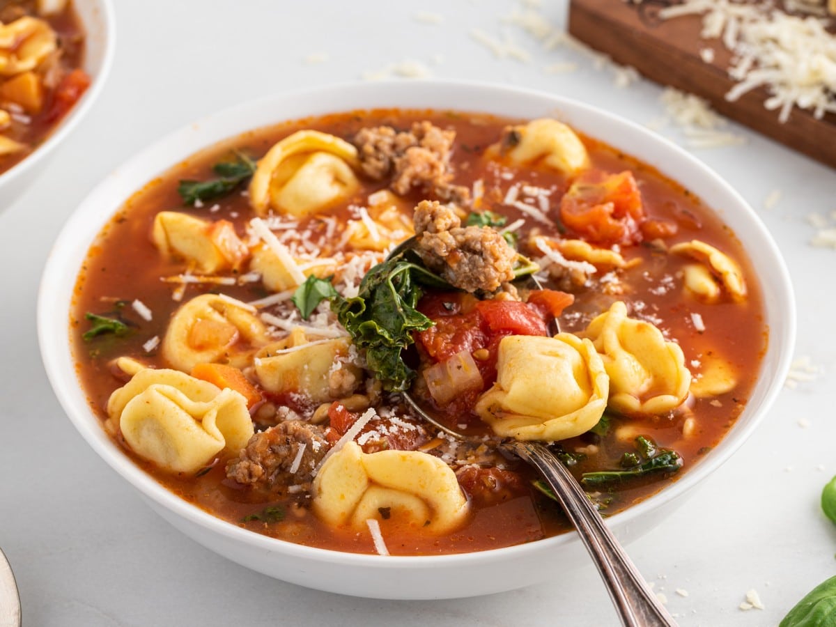 Bowl of tortellini soup topped with Parmesan cheese, with cheese tortellini, sausage, tomatoes, kale, and carrots in tomato broth.