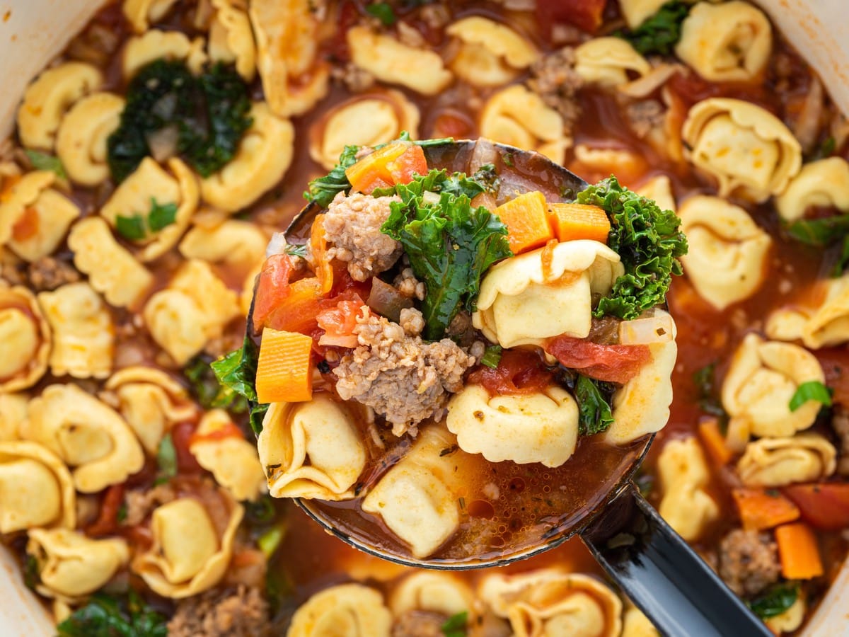 Overhead close-up of a black ladle filled with tortellini soup, showing cheese tortellini, sausage, kale, carrots, and tomatoes above the pot.