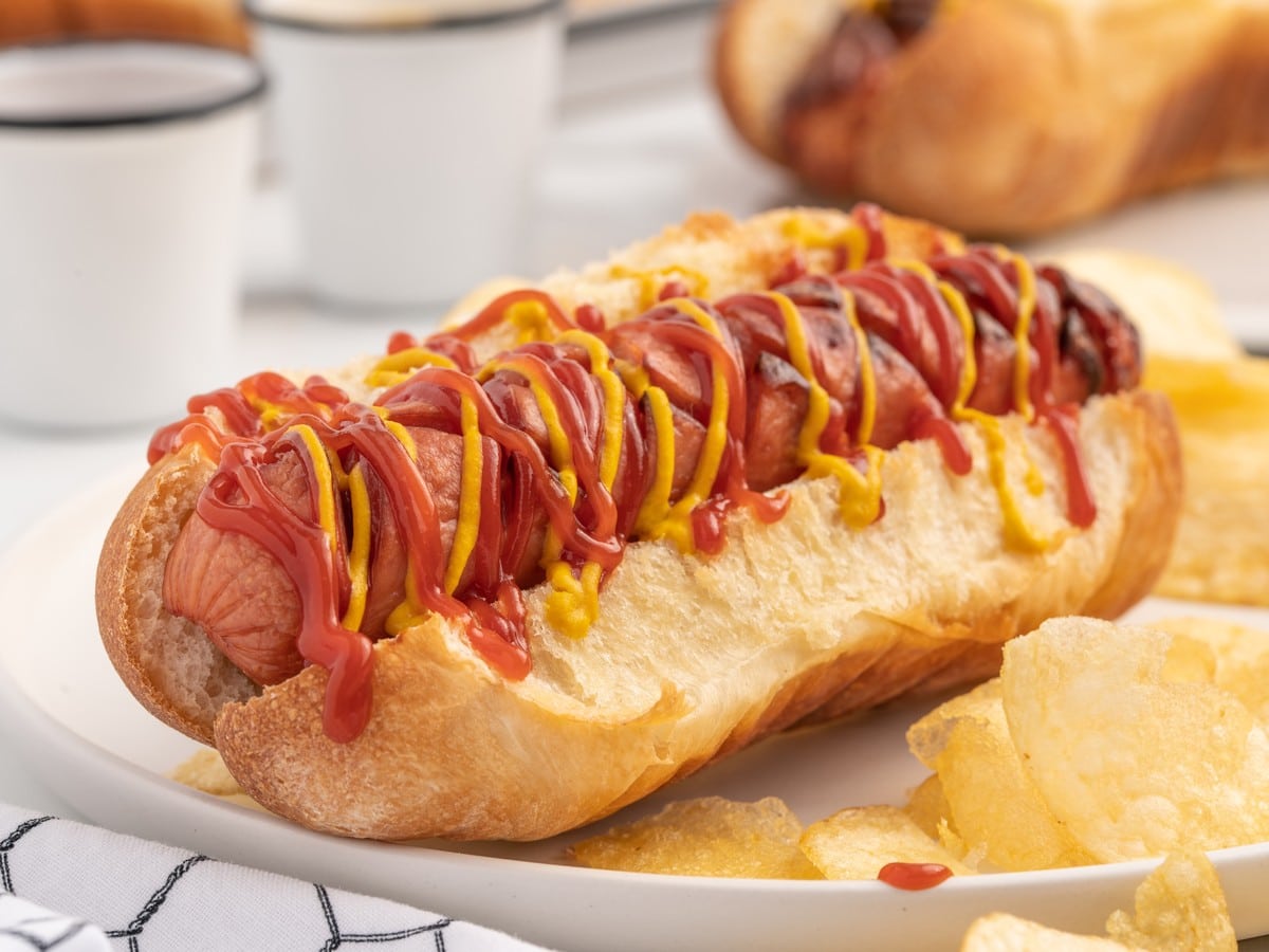 Close-up hero image of an air fryer hot dog in a toasted bun drizzled with ketchup and mustard, served on a plate with potato chips for an easy air fryer hot dog recipe