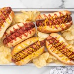 Recipe card image for air fryer hot dogs showing four toasted hot dogs in buns topped with ketchup and mustard, arranged with potato chips on a serving tray for an easy homemade air fryer hot dog recipe