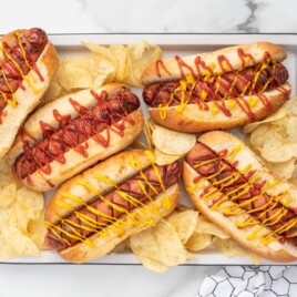 Recipe card image for air fryer hot dogs showing four toasted hot dogs in buns topped with ketchup and mustard, arranged with potato chips on a serving tray for an easy homemade air fryer hot dog recipe