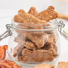 Bacon Dog Treats stored in a clear glass jar with extra bone-shaped treats and crispy bacon pieces arranged around it on a white surface.