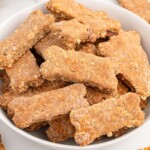 Close-up of homemade Bacon Dog Treats shaped like bones piled in a white bowl, showing a crunchy baked texture with visible oats and bacon bits.