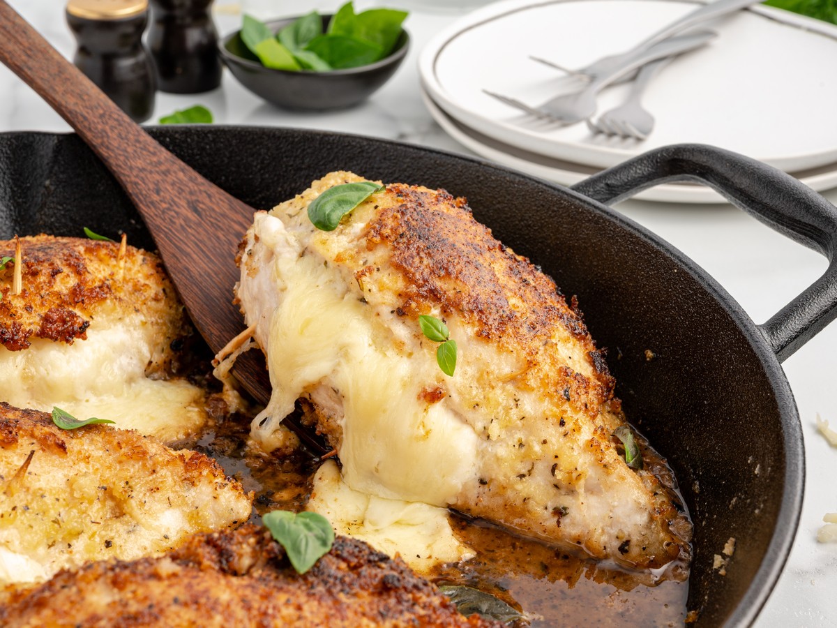 Wooden spatula lifting a cheese stuffed chicken breast from a cast iron skillet, showing melted cheese and golden baked seasoning.