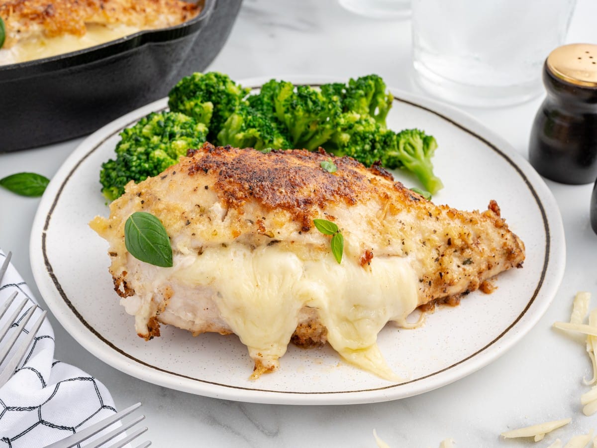 Cheese stuffed chicken breast with melted cheese spilling from the center, served on a white plate with broccoli and fresh basil.