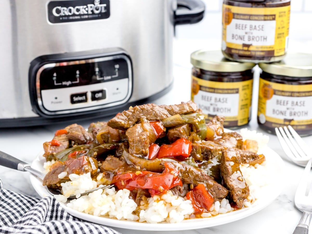 Plate of Crock Pot Pepper Steak served over rice with beef strips, peppers, onions, and sauce, styled beside a slow cooker and jars of beef base.