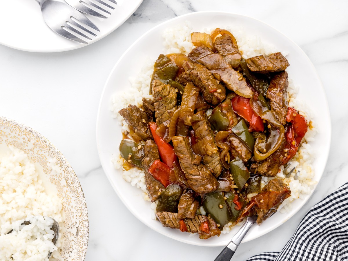 Overhead view of Crock Pot Pepper Steak served on white rice with tender beef, green and red peppers, onions, and brown sauce, with extra rice and tableware nearby.