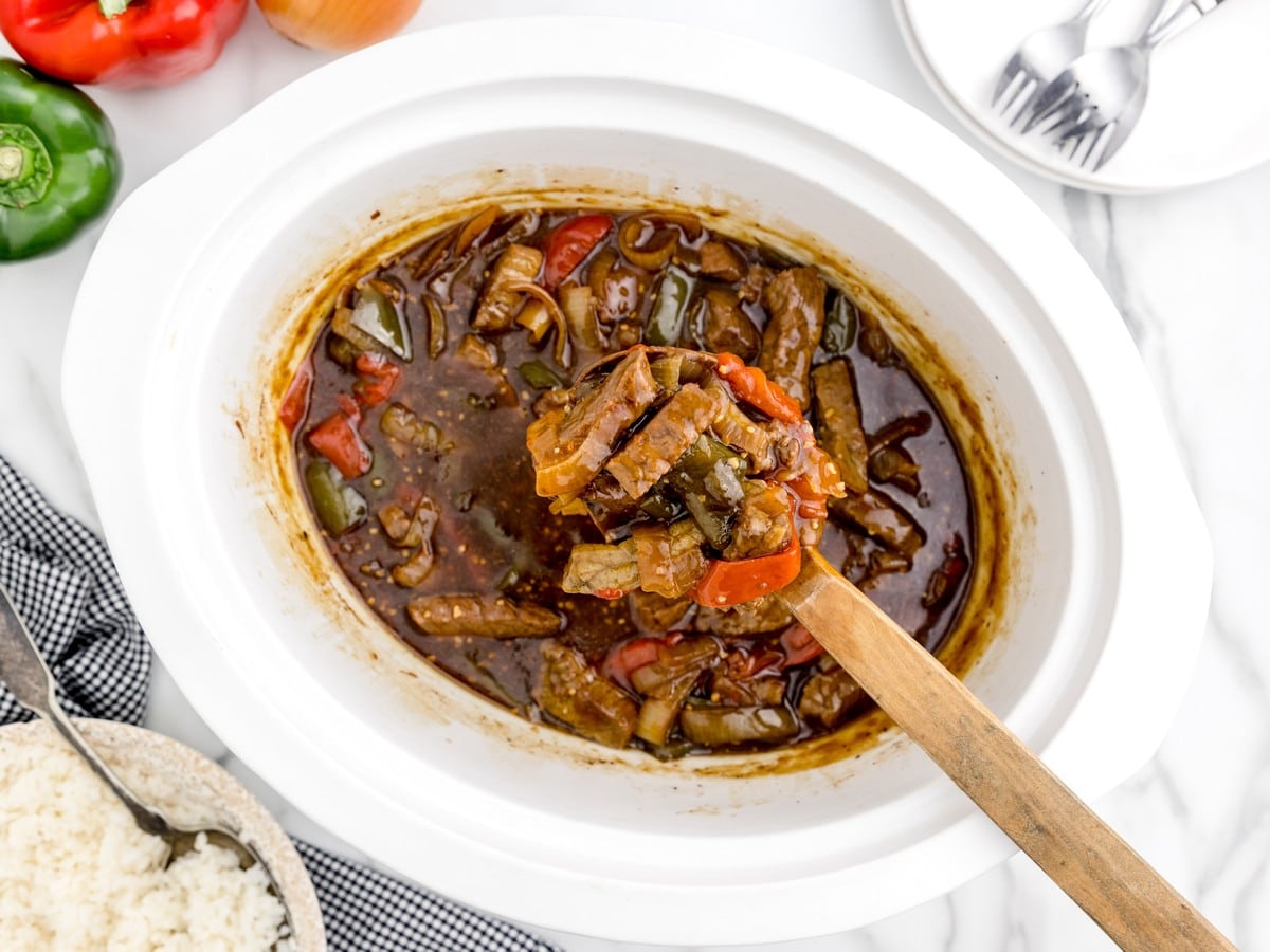 Crock Pot Pepper Steak cooking in a white slow cooker with tender beef strips, red and green bell peppers, onions, and glossy brown sauce, with a wooden spoon lifting a serving.