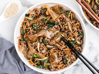 White bowl of Korean noodles with ground beef, spinach, carrots, onions, sesame seeds, and black chopsticks on a marble surface.