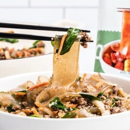 Close-up of Korean noodles with chopsticks lifting a wide glass noodle from a bowl filled with ground beef, spinach, carrots, and onions.