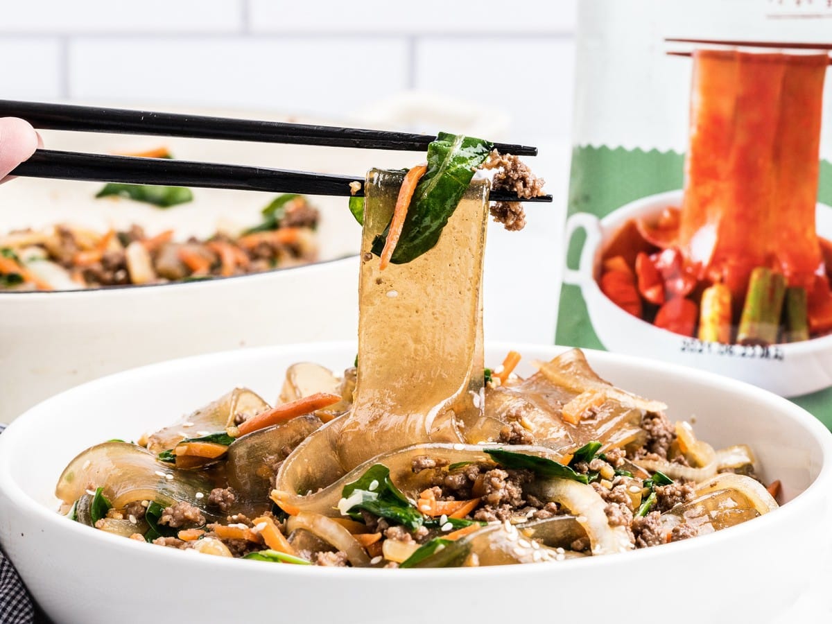 Close-up of Korean noodles with chopsticks lifting a wide glass noodle from a bowl filled with ground beef, spinach, carrots, and onions.