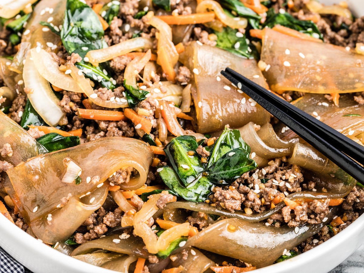 Close-up bowl of Korean noodles with glossy glass noodles, ground beef, spinach, carrots, onions, sesame seeds, and black chopsticks.