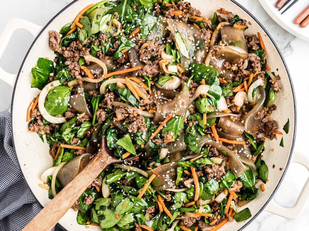 Korean noodles cooked in a large skillet with ground beef, spinach, carrots, onions, sesame seeds, and glossy wide glass noodles.