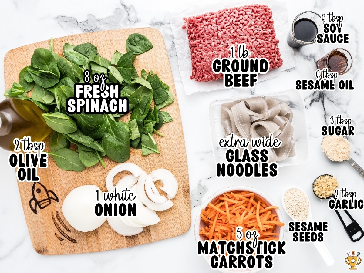 Overhead ingredient photo for Korean noodles with labeled ingredients including ground beef, Korean glass noodles, spinach, carrots, onion, soy sauce, sesame oil, garlic, sugar, olive oil, and sesame seeds.