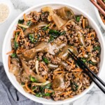 Overhead photo of Korean noodles in a white bowl with glossy Korean glass noodles, ground beef, spinach, carrots, onions, sesame seeds, and black chopsticks.