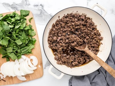 Alt text: Ground beef cooked in a white skillet with savory sauce, next to a wooden cutting board with sliced onions and fresh spinach