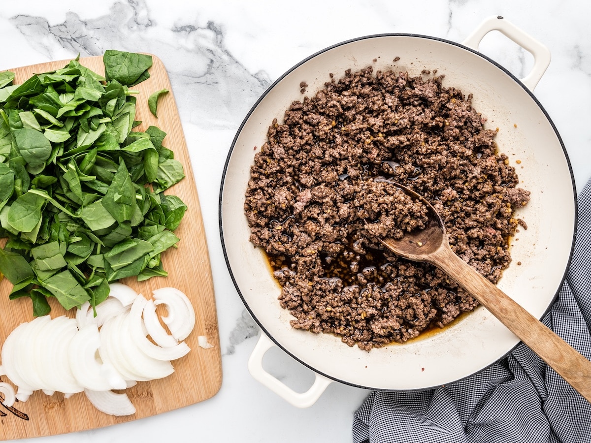 Alt text: Ground beef cooked in a white skillet with savory sauce, next to a wooden cutting board with sliced onions and fresh spinach