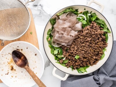 Cooked Korean glass noodles and seasoned ground beef added to a white skillet with fresh spinach, onion, and carrots