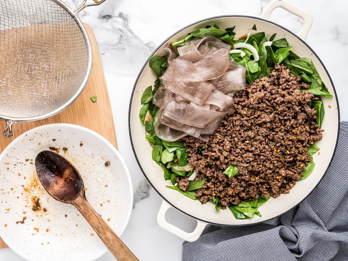 Cooked Korean glass noodles and seasoned ground beef added to a white skillet with fresh spinach, onion, and carrots