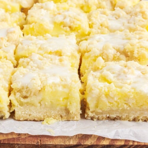 Close-up of lemon pie bars with bright lemon filling, buttery crumb topping, and sweet glaze on parchment paper over a wooden board.