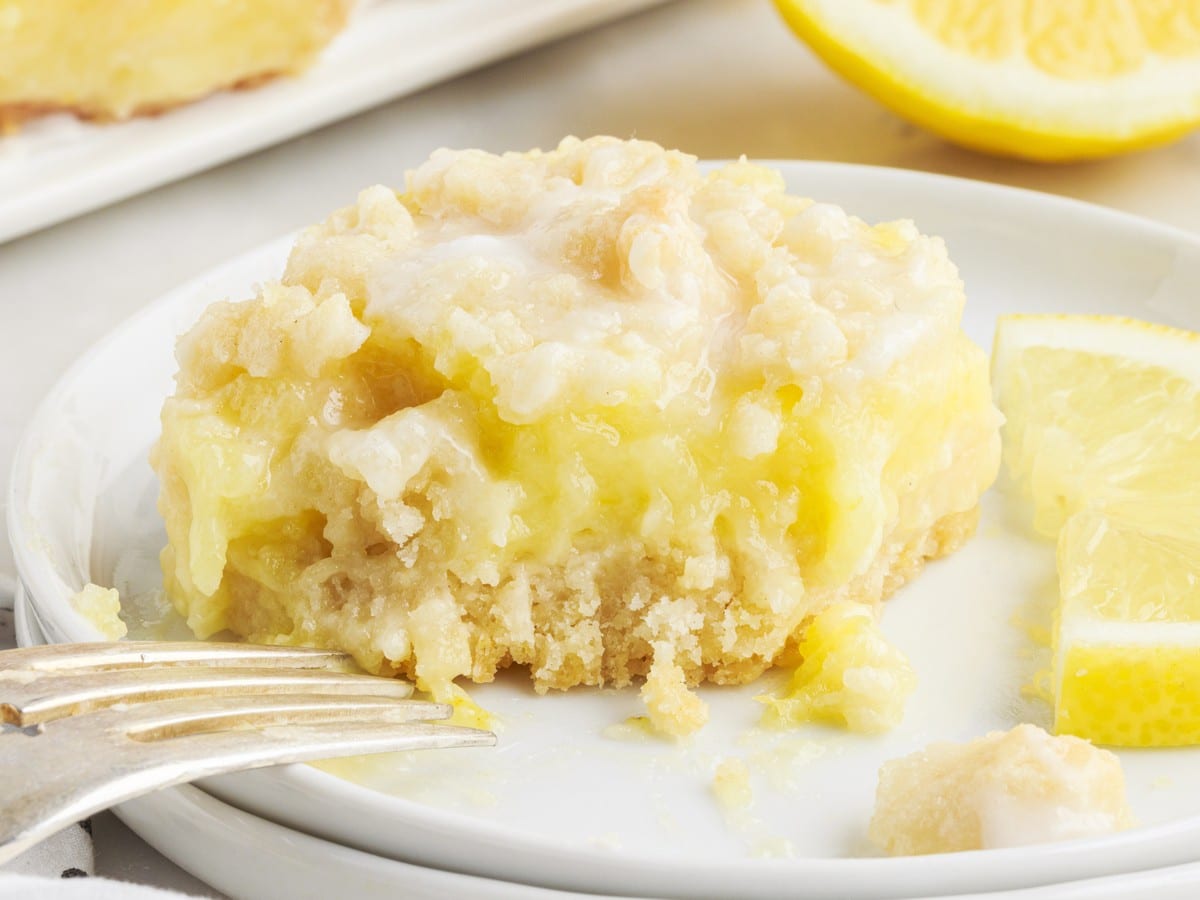 Single lemon pie bar on a white plate with creamy lemon filling, crumb topping, glaze, lemon wedges, and a fork beside it.