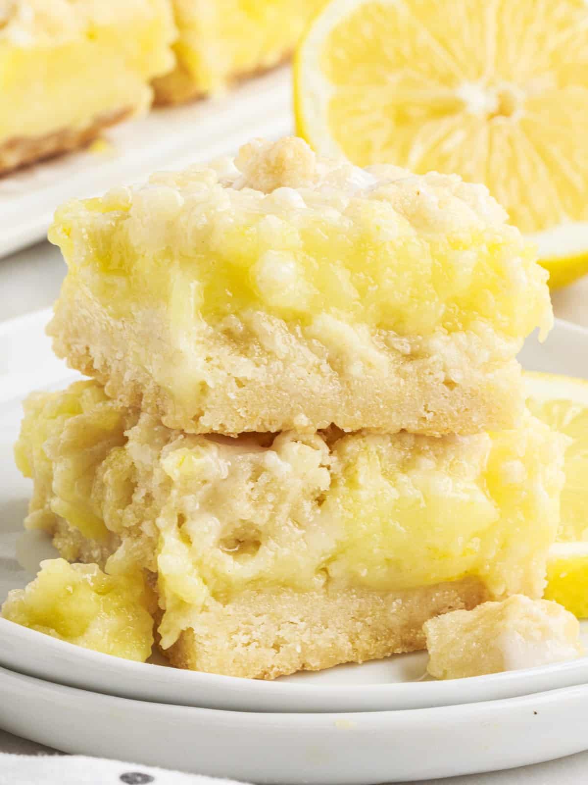 Stack of lemon pie bars on a white plate with creamy lemon filling, buttery crust, crumb topping, and fresh lemon slices in the background.