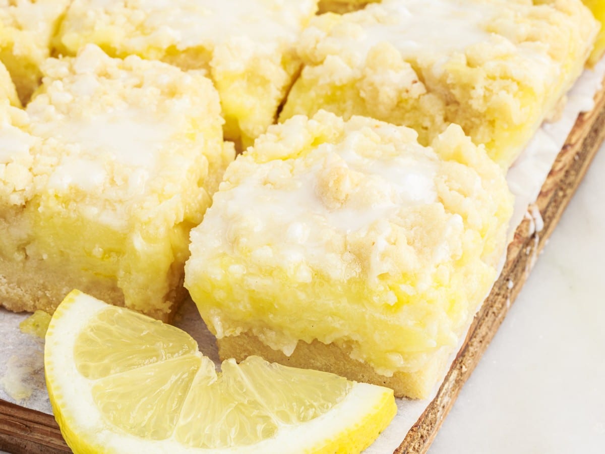 Close-up of homemade lemon pie bars with rich lemon filling, crumb topping, sweet glaze, and fresh lemon wedges on a wooden serving board.