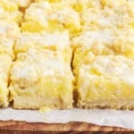 Close-up of homemade lemon pie bars with creamy lemon filling, buttery shortbread-style crust, crumb topping, and sweet glaze arranged on parchment paper over a wooden serving board.