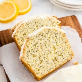Sliced lemon poppy seed bread loaf with thick lemon glaze, tender poppy seed crumb, and fresh lemons on a wooden board.