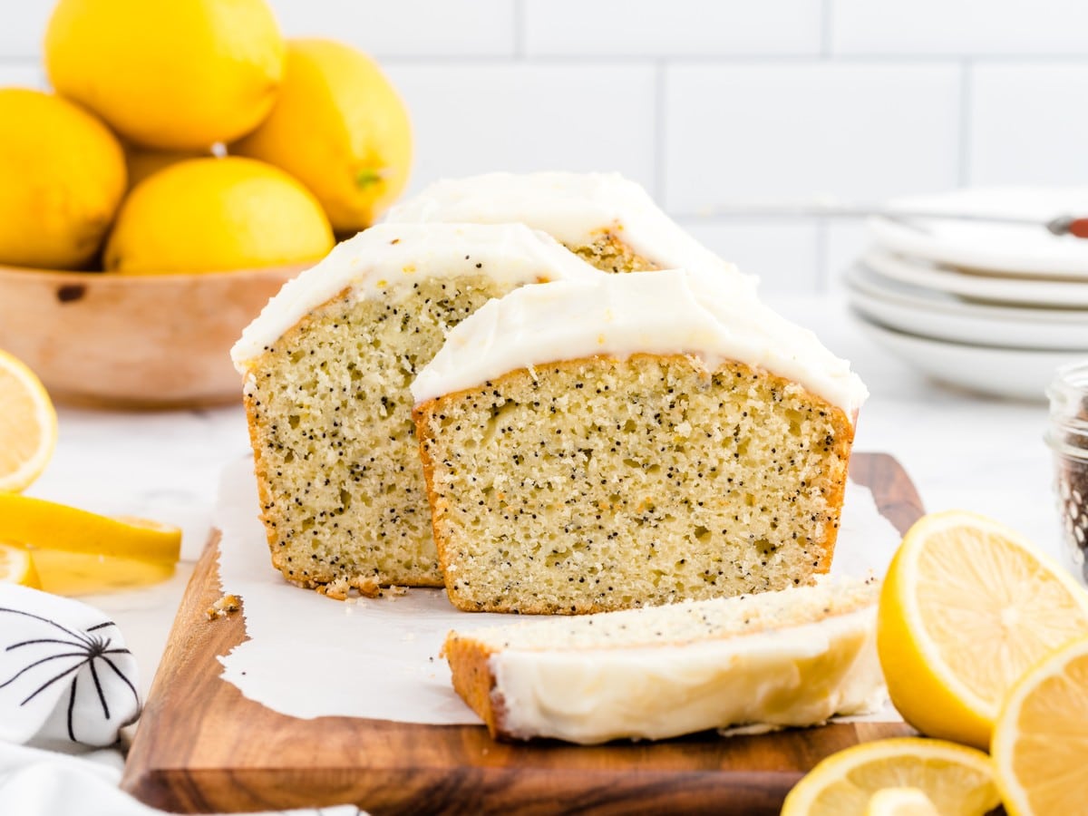 Lemon poppy seed bread loaf sliced on a wooden board with creamy glaze, fresh lemons, and a soft lemon-speckled crumb.