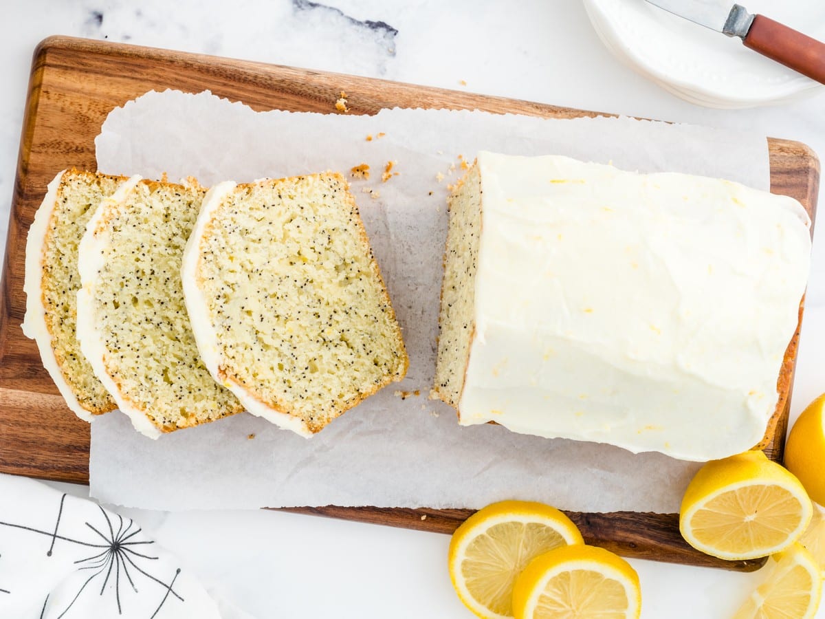 Overhead view of lemon poppy seed bread sliced on a wooden board, topped with creamy lemon glaze and served with fresh lemon slices.